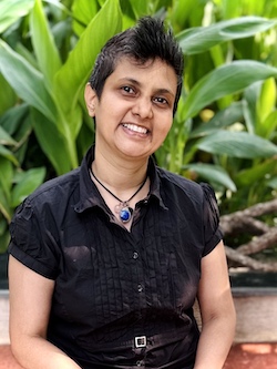 Smiling Indian lady with super short hair (mohawk), wearing a black shirt. Green leaves in the background. Chef Susmitha Veganosaurus.