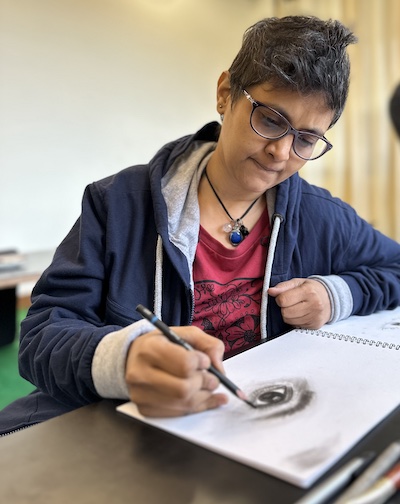 Susmitha (Indian woman, spiky short hair, wearing glasses, focused expression), doing a charcoal sketch. Dissolving the ego in deep flow.