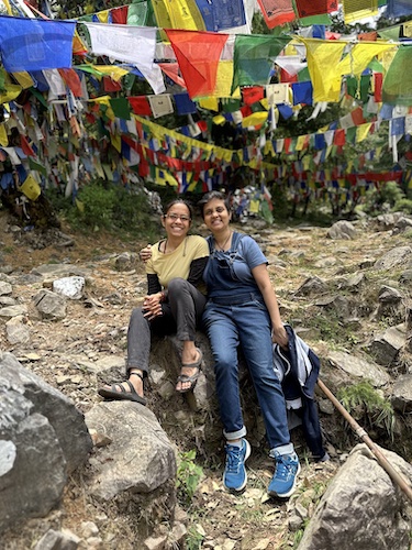 Ekta and Susmitha. Two tiny Indian women friends. Smiling wide. Sitting on a rock. Buddhist flags in the background. Location: Flag Point, Dharamkot.