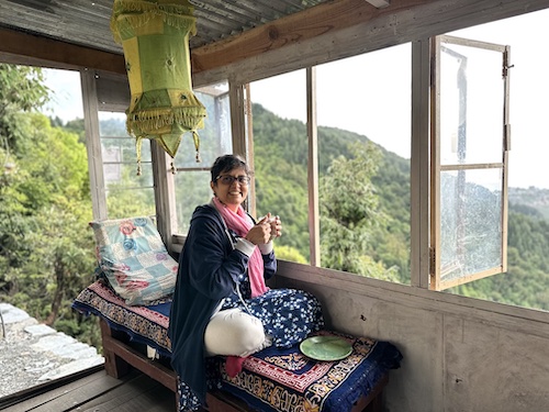 Susmitha, short haired Indian lady with glasses. Sitting cross legged on a bench, smiling at the camera. Sky and mountains outside the window.