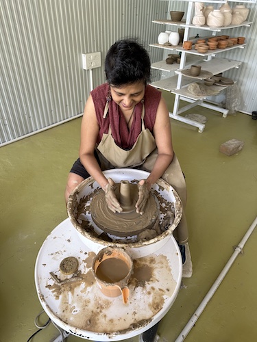 Susmitha, short haired Indian lady, intently looking down an working with terracotta on a pottery wheel. Location: Dharamkot Studio
