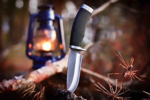 Close up of knife stuck in the earth, twigs on the side, old style lamp in the background.