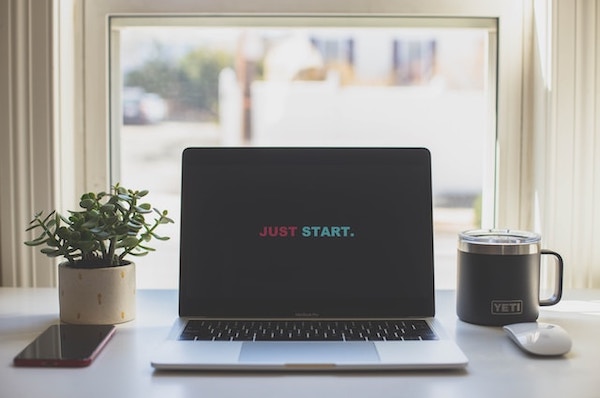 Start with what you have. Photo of an open laptop on a desk with the text "JUST START" on the screen. Plant, mug and mouse on the sides.
