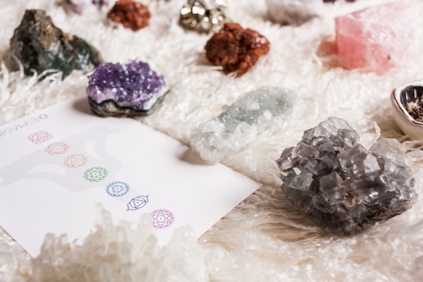 Different coloured crystals on a soft surface, next to a white sheet of paper with the seven chakras drawn on it. Chakra Harmony Meditation.