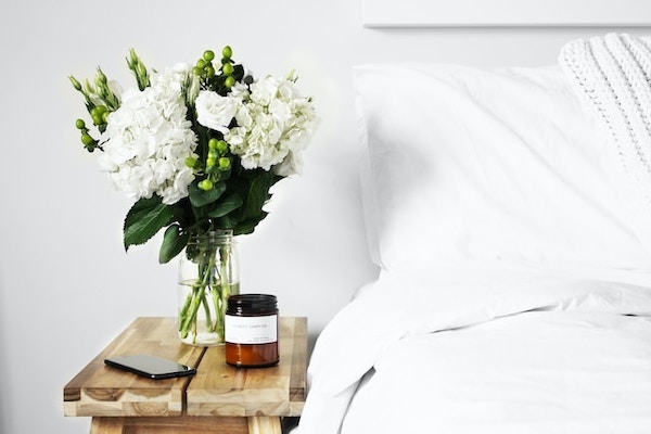 Partial view of bed and pillow with focus on the bedside wooden table. The table has a soy wax candle, glass vase with white flowers and green leaves, and a mobile phone. Morning habits.