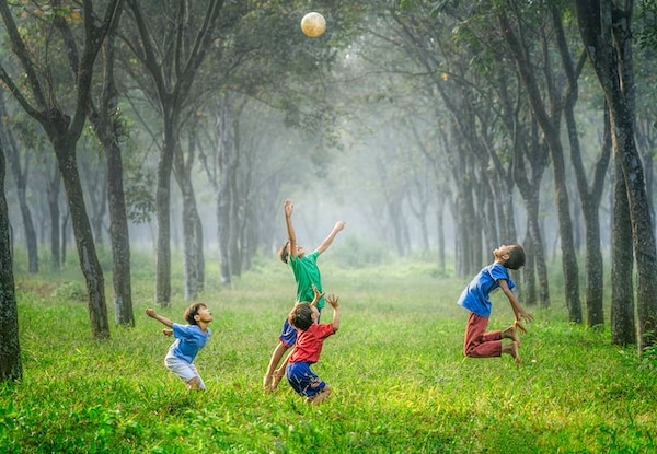 Fresh green grass grown wild. Trees in the background. Four kids happily playing with a ball with no agenda. They look delighted!
