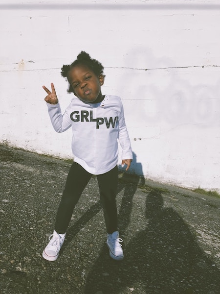 Cute little black girl in black pants and white full sleeved tee, standing with her feel apart like a boss and doing the peace sign.