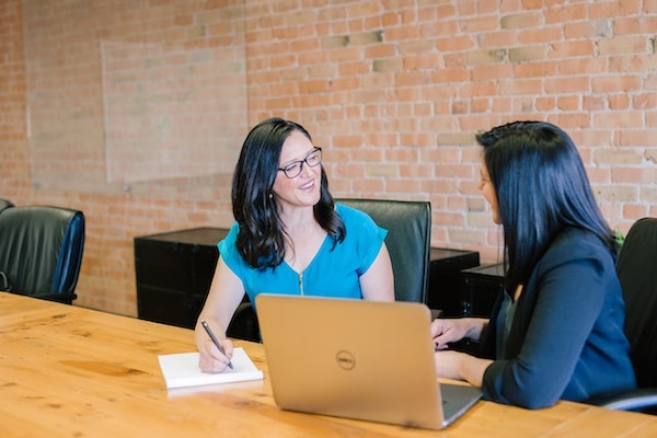 Two women with dark hair sitting next to each other at a table, in what seems to be a coaching session. One is talking (we can't see her face), the other (wearing glasses) is smiling, listening, and taking notes. Table has an open macbook on it.