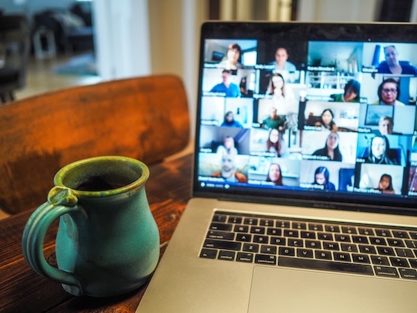 Macbook Pro open on desktop with zoom session showing group of people on it. Blurred faces. Aqua gree ceramic mug next to the laptop.