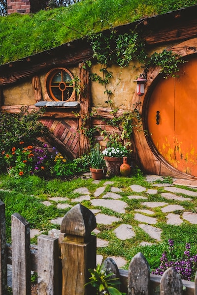 Outside a fairy tale style cottage with low roof, round door and window. Picket fence and little garden.