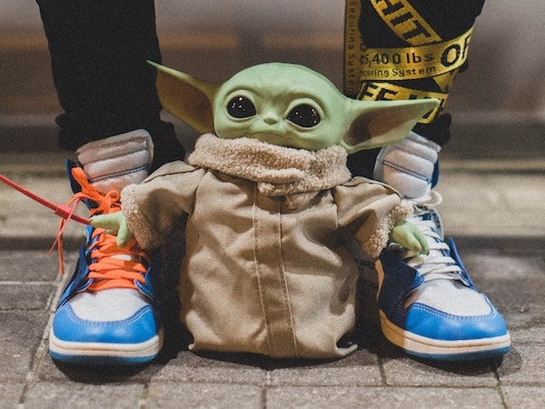 Pair of legs wearing sneakers with two different coloured laces. Adorable Grogu toy standing between the feet.