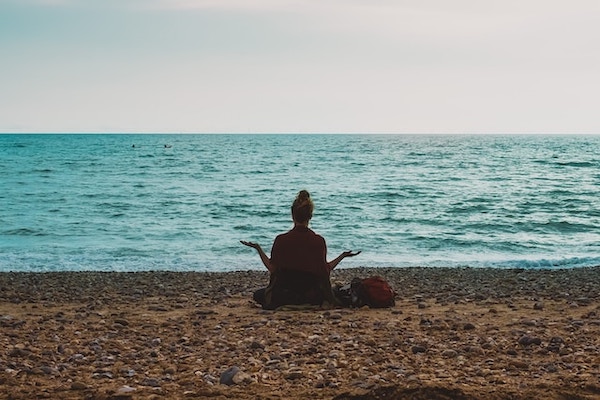 Woman sitting on a beach with her back facing us. She's sitting cross legged in a position of meditation. Arms raised as if she's setting boundaries and saying no with love and firmness.