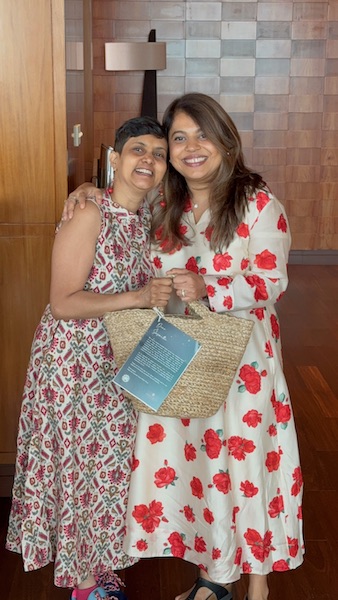 Two Indian ladies in red printed dresses. Side hugging and smiling wide at the camera. Part of a community preview event for Namu Recommends Vegan Market