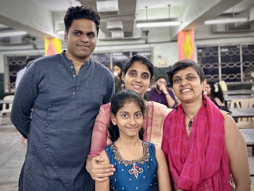 Mupaal Vegan family with Veganosaurus. Indian man, two women, one young girl. All dressed up in bright traditional wear. Standing together and smiling at camera.
