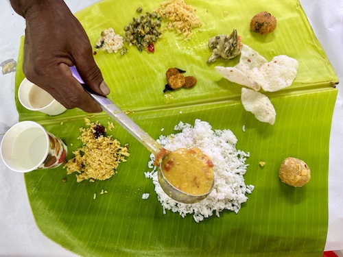 Vegan South Indian Lunch spread, served traditionally on banana leaf. Hand holding serving spoon with dal over the white rice in the full leaf.