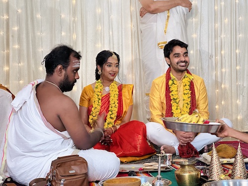 South Indian Wedding. Shaadu and Venky. Bride in orange saree and groom in yellow kurta and while pants seated at floor level in padmasana. Priest on one side doing his thing.