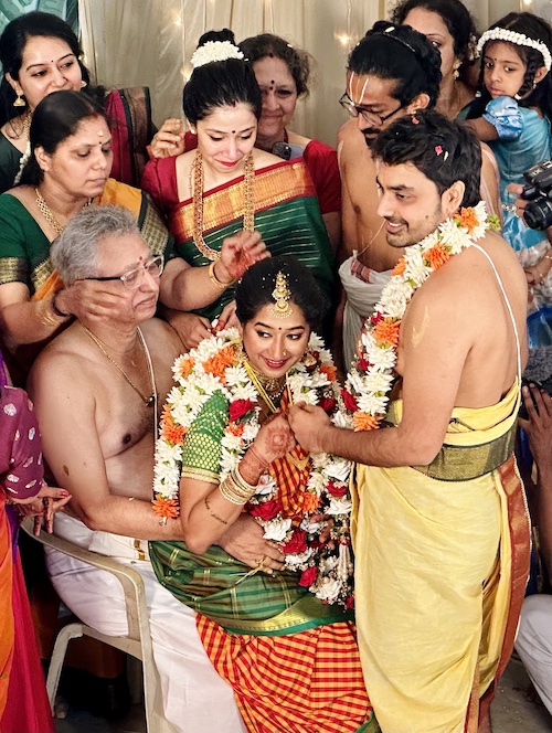 South Indian Wedding. Shasvathi Siva and Venkataraghavan. Bride sitting and smiling, looking to one side. Groom standing in front of her, holding her hands and also looking to the side. People surround them looking happy. All dressed in bring traditional clothes.