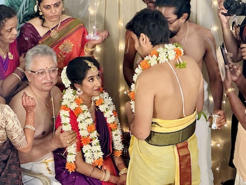 South Indian Wedding. Shasvathi Siva and Venkataraghavan. Bride sitting on older gentleman's lap, looking up with a smile at the groom who is standing and facing her. Both wearing garlands.
