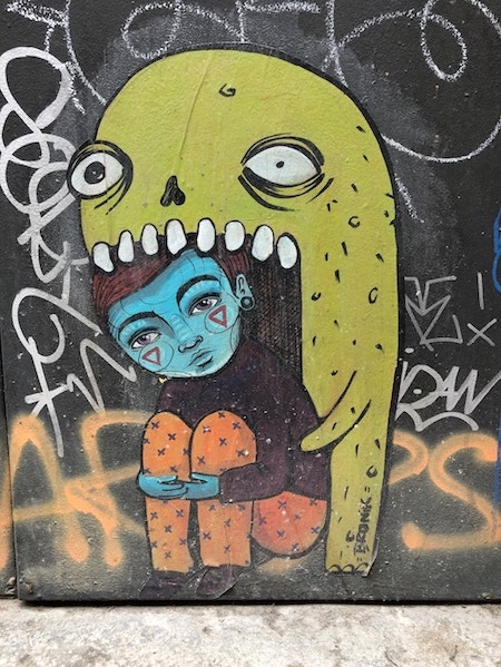 Street art of green monster trying to eat a little human being who's sitting on the floor with their legs drawn in to their chest, their arms wrapped around their shins.