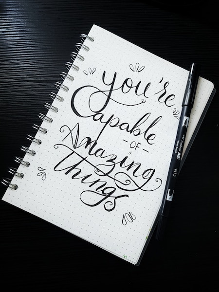 Ruled, spiralbound notebook on a black table with pen on it. Beautiful cursive writing on the page: You're capable of amazing things.
