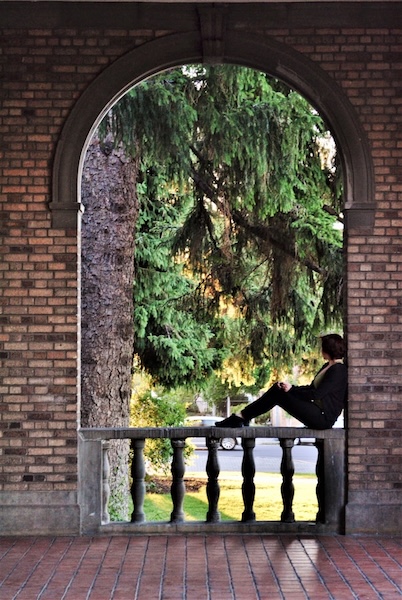 Person sitting on a bannister under a brick wall arch, staring off calmly into nature far away. Moment of peace and ease.
