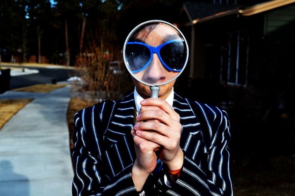 Person wearing blue sunglasses, holding up a magnifying glass to their face so we can see an enlarged version of their nose and upper part of face.
