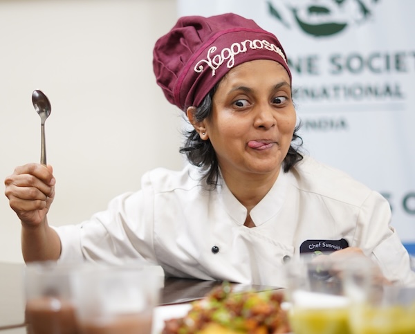 Chef Susmitha Veganosaurus, wearing a white chef coat and maroon chef hat. Ready to dig into a vegan meal after a plant based cooking workshop at a culinary college in Bangalore, India.
