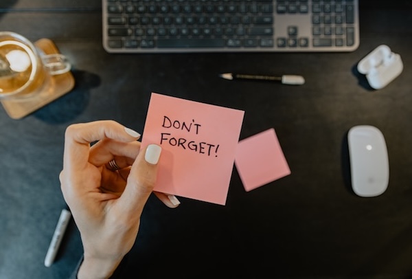 Hand with nails painted white, holding up a pink square piece of paper with words "DON'T FORGET". Make use of reminders, calendar and alarom to reduce your mental load.