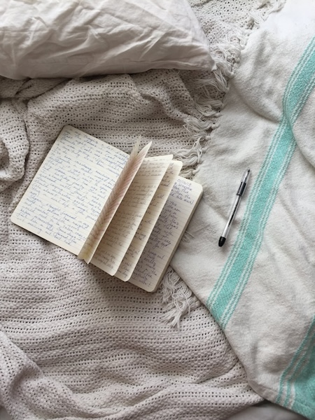 Open notebook and pen strewn casually on a bed with blankets. There's flowing writing in the book. Writing is a low maintenance creative activity.