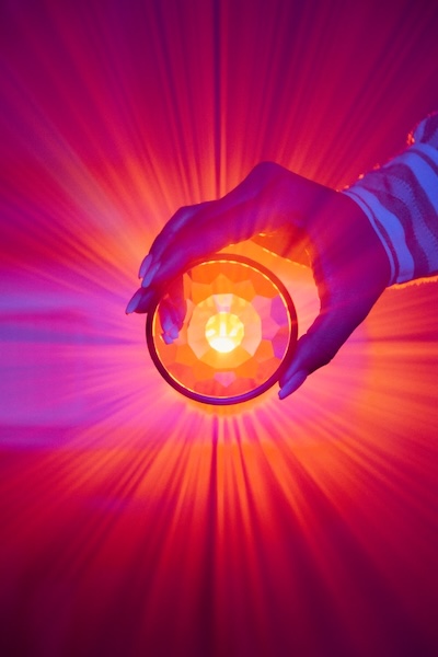 Bright and dark photo with lots of shades of glowing reds and pinks. Hand with long fingernails, holding something like a glass crystal to light. Light shining through it and beaming out. With a regular free-writing practice, you can channel spirit onto the page.