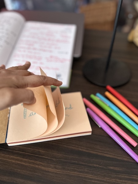 Cocolourful sketch pens and a notepad on a table, pages being flipped by a hand.