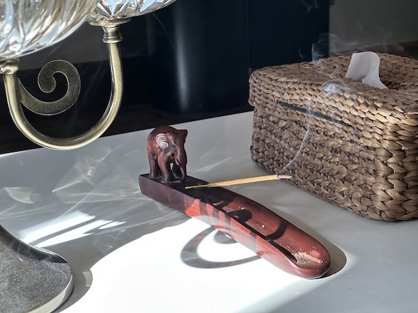 Wooden insence holder with a carved elephant on one end. Incense wth swirls of smoke rising from it. Light and shadow play on the table it's kept on. Joy lives in the little moments and small details.