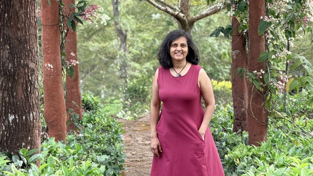 Susmitha Veganosaurus. Vegan chef, meditation teacher, business mentor, coach for multi-passionate entrepreneurs. In a maroon dress, smiling at camera, one hand in pocket.